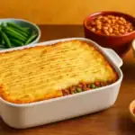 Cottage pie in a white dish surrounded by green beans, baked beans, and coleslaw on a vibrant wooden kitchen table.