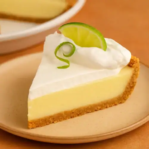 A close-up slice of key lime pie with a graham cracker crust, whipped cream, and fresh lime garnish on a warm-toned plate.