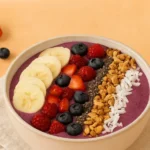 Smoothie bowl topped with banana slices, berries, granola, chia seeds, and coconut flakes on a warm background