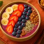 Close-up of a vibrant smoothie bowl topped with banana, strawberries, blueberries, granola, and hemp seeds on a wooden table.