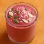 Glass of pink smoothie topped with pumpkin seeds, sliced almonds, and shredded coconut on a warm orange background.