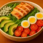 Cobb salad with grilled chicken, avocado, blue cheese, boiled eggs, and cherry tomatoes in a white bowl on a wooden table