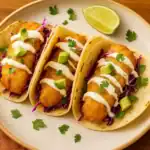 Close-up of three crispy Baja fish tacos topped with lime crema, cabbage slaw, avocado, and cilantro on a plate.