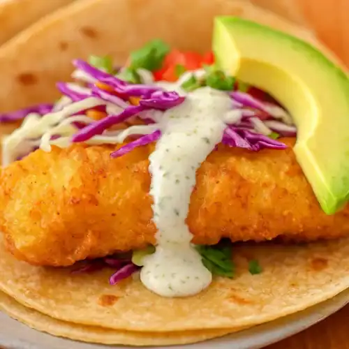 Close-up of a crispy Baja fish taco with cabbage, lime crema, and avocado on a corn tortilla.