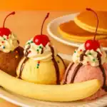 Classic homemade banana split with scoops of chocolate, vanilla, and strawberry ice cream topped with whipped cream, cherries, and served with pancakes in the background.