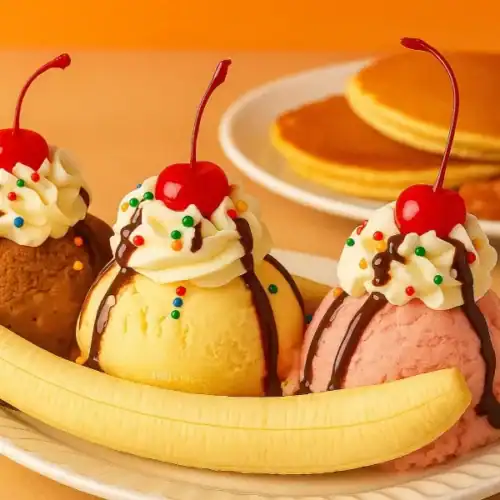 Classic homemade banana split with scoops of chocolate, vanilla, and strawberry ice cream topped with whipped cream, cherries, and served with pancakes in the background.