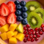 Smoothie bowl topped with sliced strawberries, blueberries, kiwi slices, mango chunks, and pomegranate seeds.