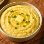Bowl of creamy potato grits topped with butter and green onions, with a spoon on the side