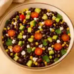 A bowl of vibrant black bean and corn salad with cherry tomatoes, diced cucumber, corn, feta, and fresh cilantro.