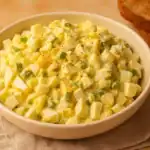Close-up of classic egg salad in a cream bowl, served with chopped green onions and a side of toasted bread.