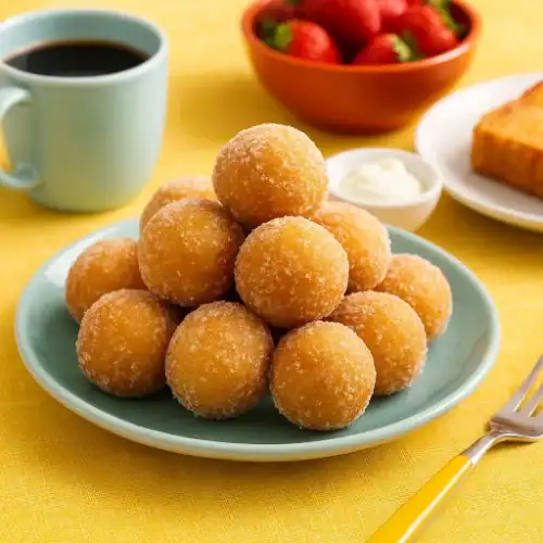 A plate of golden, sugar-coated 3-ingredient donut holes served with coffee and strawberries on a vibrant summer breakfast table.