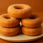 Stack of golden-brown buttermilk donuts on a beige plate with a warm orange background.