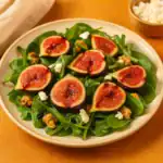 Close-up of a fig salad with fresh spinach, goat cheese, and walnuts on a beige plate with a warm orange background