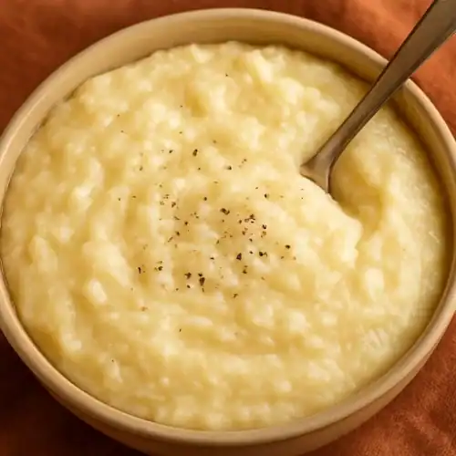 Close-up of creamy potato grits in a bowl with a spoon and a sprinkle of black pepper