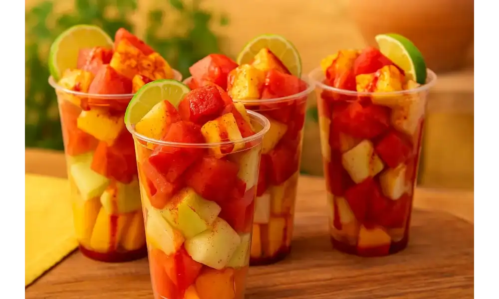Four colorful Mexican fruit cups filled with mango, watermelon, pineapple, and cucumber, topped with chili powder and lime wedges.