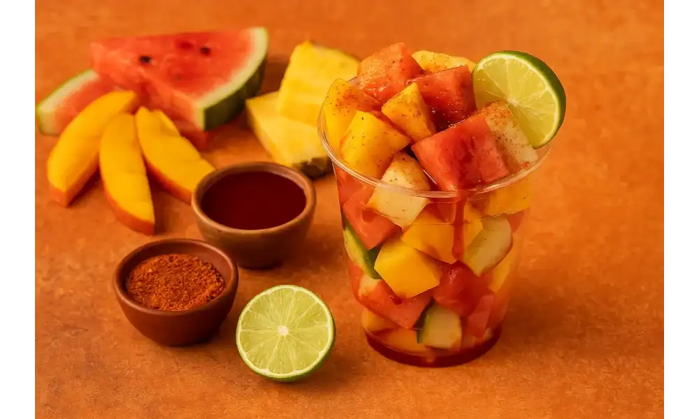 A single Mexican fruit cup filled with mango, watermelon, jicama, and cucumber, topped with chili powder and lime, surrounded by fresh ingredients.