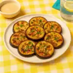 Mediterranean roasted eggplant slices on a white plate, served on a yellow checkered tablecloth with lemon water and tahini dip.
