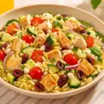 A bowl of Mediterranean orzo chicken salad with grilled chicken, cherry tomatoes, cucumbers, olives, spinach, red onion, and feta cheese served on a summer table.