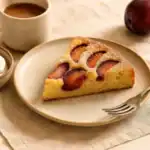 A slice of homemade plum cake on a plate served with coffee in a cozy breakfast setting.