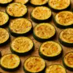 Close-up of golden roasted zucchini slices on parchment paper, seasoned with herbs and spices.