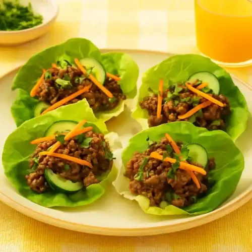 Beef and lettuce wraps served on a plate with cucumber slices, carrots, and cilantro in a bright summer setting with orange juice.