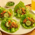 Plate of beef and lettuce wraps filled with ground beef, cucumber slices, carrots, cilantro, and chili flakes, served outdoors with a glass of juice.