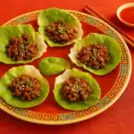 Chinese beef lettuce wraps on a red patterned plate, garnished with scallions and sesame seeds, served with chopsticks and dipping sauce on a red table setting.