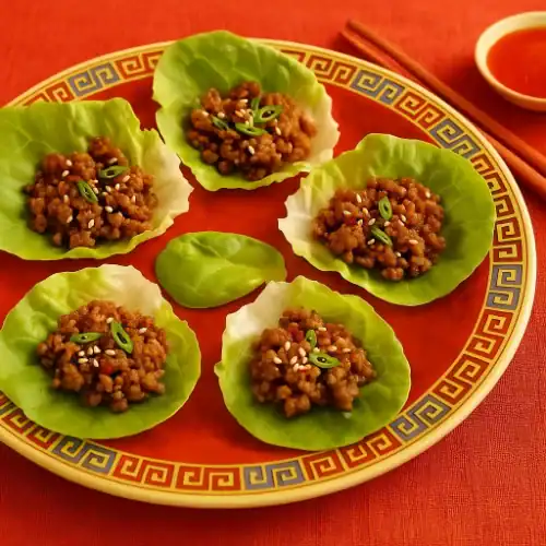 Chinese beef lettuce wraps on a red patterned plate, garnished with scallions and sesame seeds, served with chopsticks and dipping sauce on a red table setting.