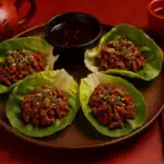 Chinese beef lettuce wraps served on a brown plate with scallions and sesame seeds, styled with chopsticks, chili sauce, and tea on a red Chinese table setting.