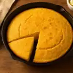 Freshly baked golden Jiffy cornbread in a cast-iron skillet with a slice cut out, served on a wooden table.