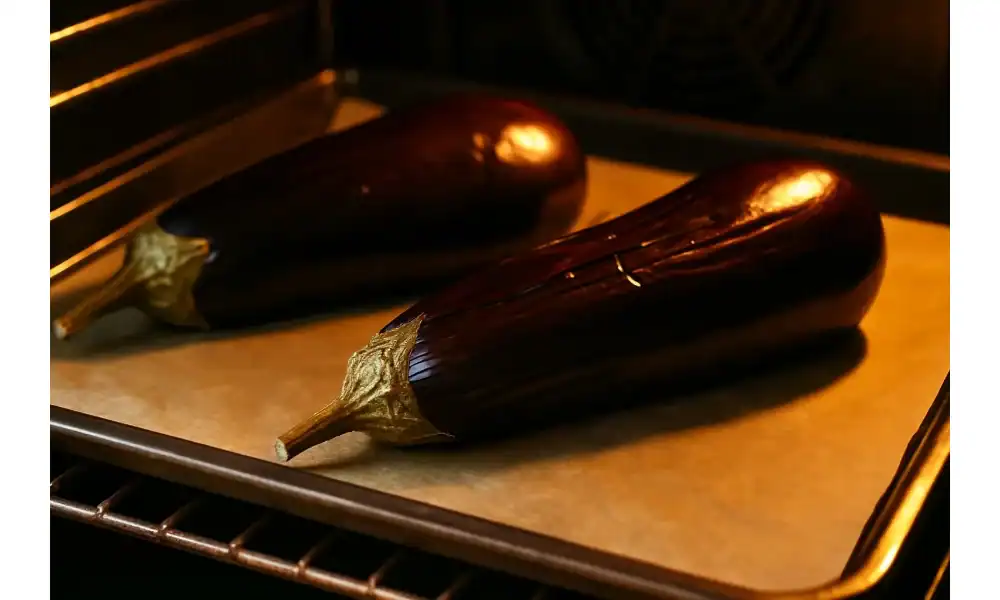 Two whole eggplants roasting on a parchment-lined baking sheet in a warm-lit oven.
