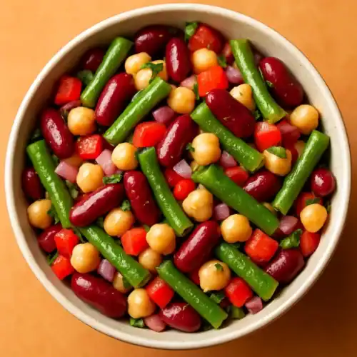 Overhead close-up of a bowl of three bean salad—kidney beans, chickpeas, and green beans with diced red pepper, red onion, and parsley in a tangy vinaigrette.