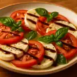 Close-up of sliced tomatoes and fresh mozzarella arranged on a ceramic plate with basil leaves, extra-virgin olive oil, and a balsamic glaze drizzle on a wooden table.