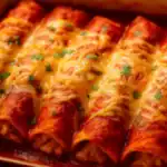 Close-up of cheesy chicken enchiladas covered in red enchilada sauce and melted Jack–cheddar, sprinkled with cilantro in a baking dish.