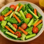 Bowl of quick finger salad with romaine, cucumber, celery and carrot sticks plus cherry tomatoes, shown on a warm wooden table with vinaigrette in the background.