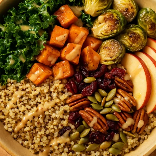 Close-up of a colorful fall harvest grain bowl with quinoa, roasted sweet potatoes, kale, Brussels sprouts, apples, cranberries, pecans, and pumpkin seeds drizzled with maple-tahini dressing.