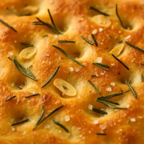 Close-up of golden garlic rosemary focaccia showing crispy crust, fresh rosemary sprigs, thin garlic slices, and flakes of sea salt under warm light.