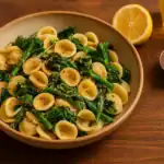 A bowl of vegan orecchiette pasta with broccoli rabe, drizzled with olive oil, and sprinkled with red pepper flakes, lemon zest, and breadcrumbs, served on a wooden table in warm light.