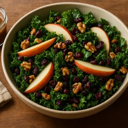 Bowl of kale apple balsamic salad with sliced apples, walnuts, and cranberries beside a jar of vinaigrette on a wooden table.
