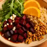 Close-up of roasted beet cubes served with quinoa, arugula, orange slices, walnuts, and crumbled feta on a beige plate.