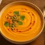 Bowl of creamy sweet potato coconut soup with chili-oil swirl, pumpkin seeds, cilantro, toasted coconut, and a silver spoon on a wooden table.