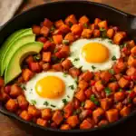 Cast-iron skillet of sweet potato hash with two sunny-side-up eggs, diced peppers and onions, avocado slices, and fresh herbs.