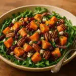 A plate of fall sweet potato salad with roasted sweet potatoes, cranberries, pecans, feta, and arugula on a rustic wooden table.
