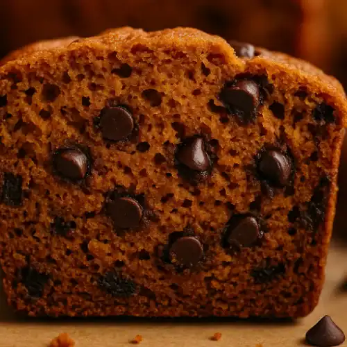 Close-up of a slice of vegan pumpkin chocolate chip bread showing moist texture and melted chocolate chips under warm lighting.