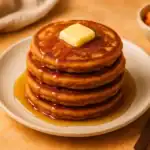 Stack of golden vegan sweet potato pancakes topped with a pat of vegan butter and drizzled with maple syrup on a ceramic plate.
